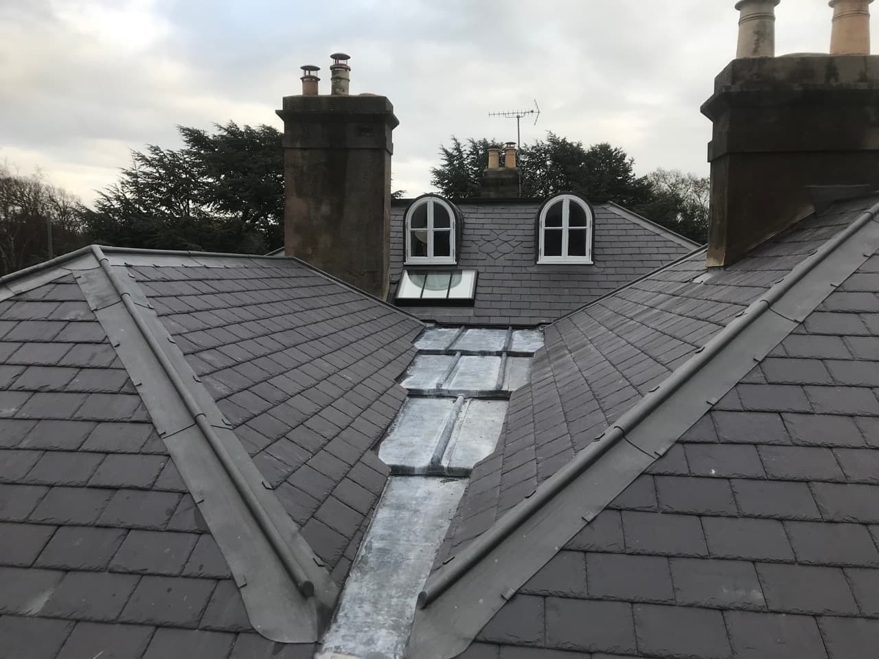 Grey slate roof with a central lead-lined valley, arched dormer windows and stone chimneys